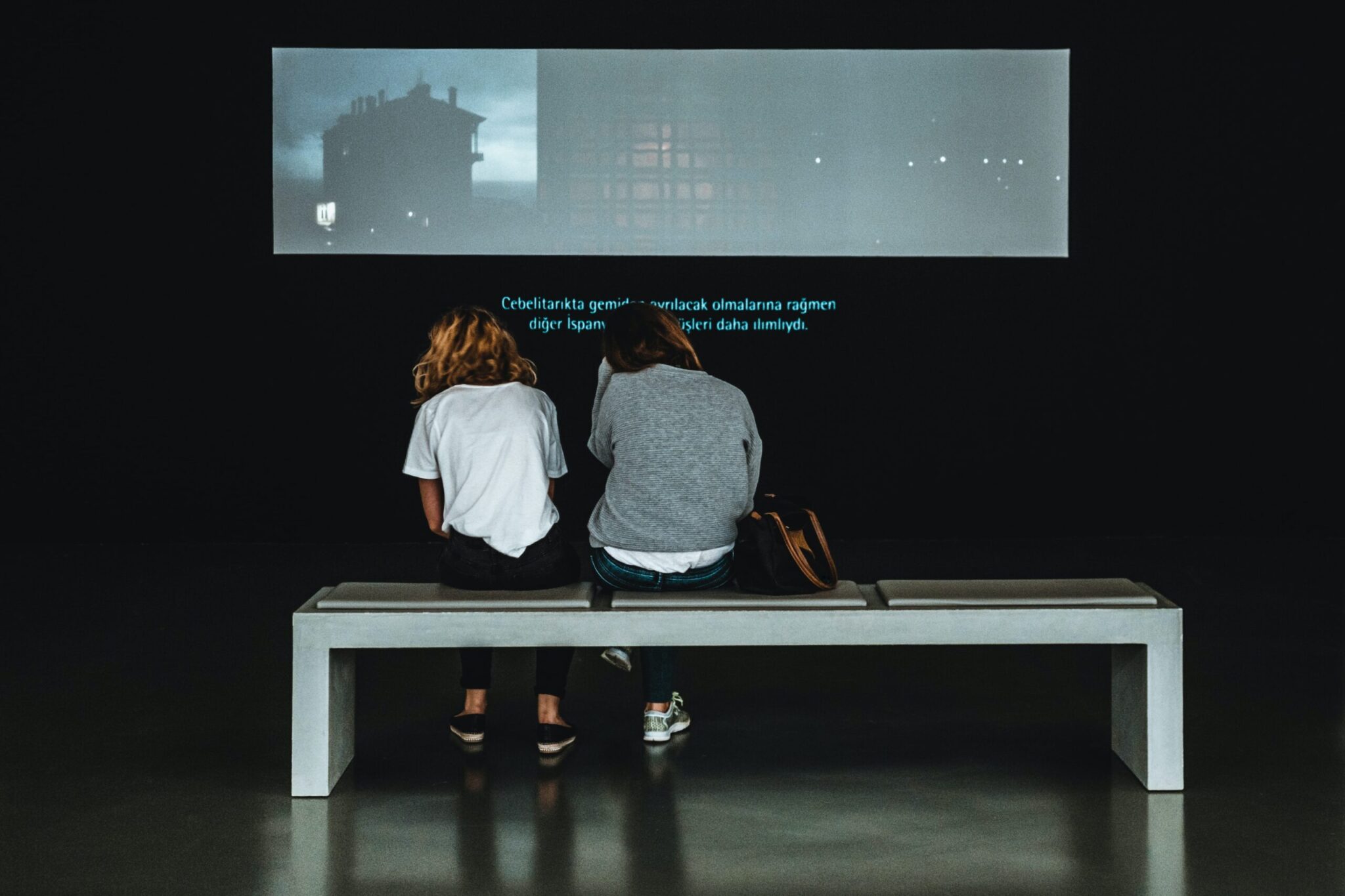 Två unga kvinnor sitter på en bänk i en mörklagd utställningslokal och ser på en projektion på en vägg.