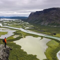 En person fjällvandrar och står i kanten av en stor dal där det rinner vatten. Rapadalen