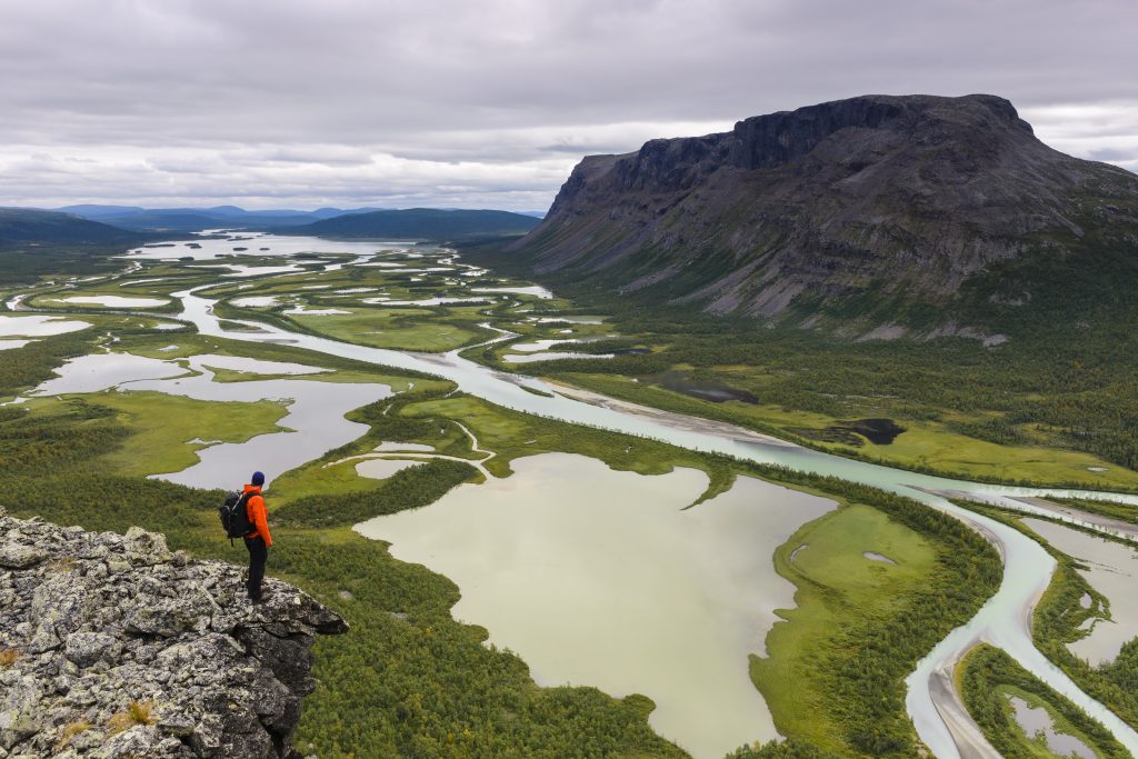 En person fjällvandrar och står i kanten av en stor dal där det rinner vatten. Rapadalen