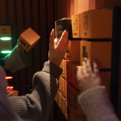 Händer från tre olika personer samlas kring och lyfter ut små orange boxar i en utställning