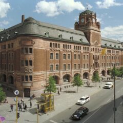 En stor äldre tegelbyggnad med klocktorn, med en gata i förgrunden.