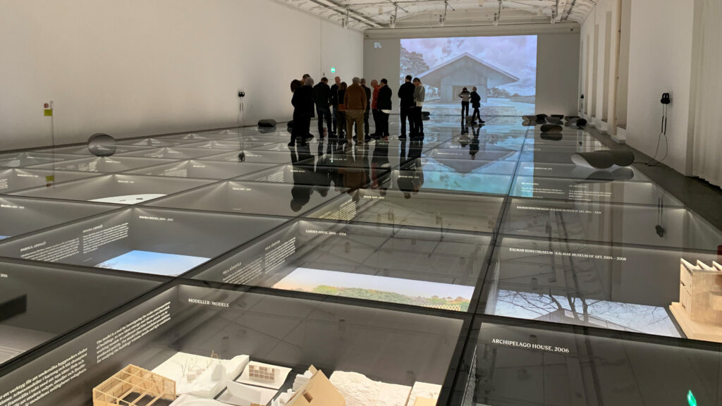 En utställningssal på Arkdes med montrar i golvet, en grupp människor står en bit bort.