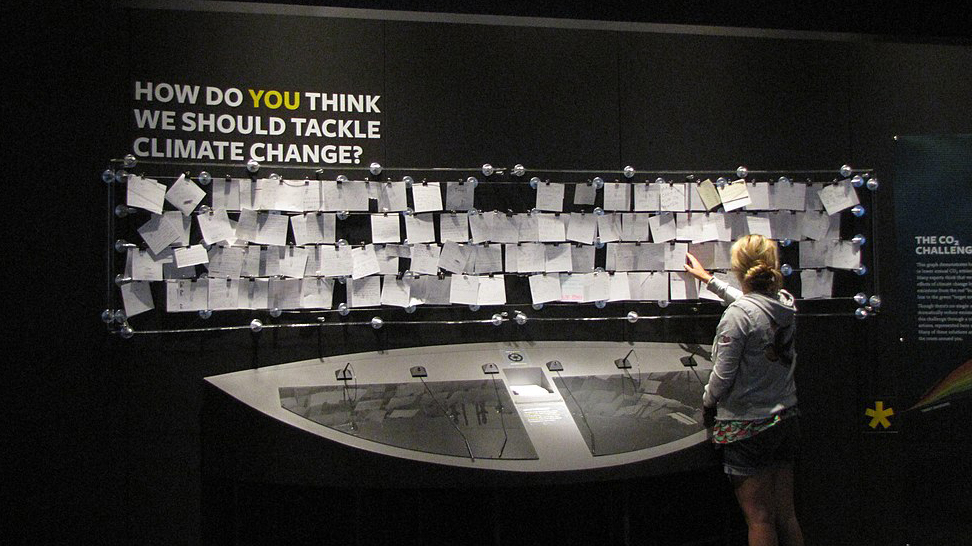 En utställningssal på Field Museum i Chicago med texten HOW DO YOU THINK WE SHOULD TACKLE CLIMATE CHANGE på väggen, under sitter lappar med förslag. En kvinna står och tittar på en av lapparna.