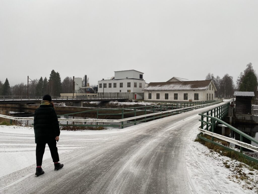 Bro som leder fram till några äldre industribyggnader. I förgrunden går en person mot bron.