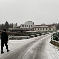 Bilden visar en bro som leder fram till några äldre industribyggnader. I förgrunden går en person mot bron.