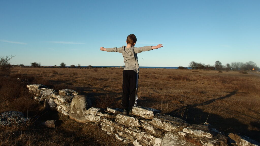 Ett barn står med utfällda armar på en mur och blickar ut över landskapet.