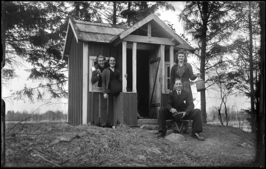 Bilden visar en lekstuga. Svartvitt foto. Fyra glada vuxna på farstun tittar in i kameran.