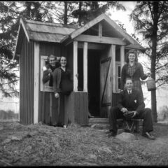 Bilden visar en lekstuga. Svartvitt foto. Fyra glada vuxna på farstun tittar in i kameran.