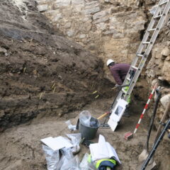 På bilden syns ett djupt schakt med en stege för att komma ner. Nere i botten står en arkeolog som dokumenterar kulturlagren. Längst fram i bild syns provpåsar och hjälpmedel för inmätning.