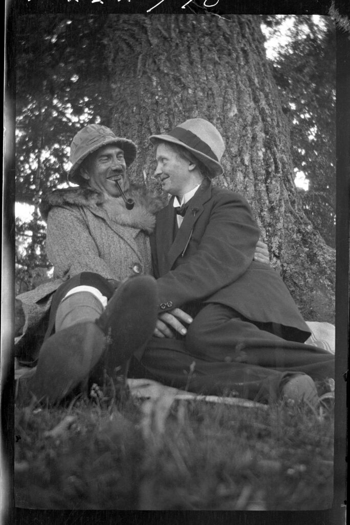 Skrattande man och kvinna som halvligger mot en trädstam. Båda har hatt, kvinnan har manskläder och mannen har tropikhatt och en pipa i munnen.