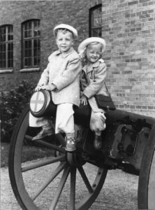 Två barn, en pojke och en flicka, klädda i kappa och hatt. Sitter på röret till en gammal kanon. Med tegelvägg i bakgrunden.