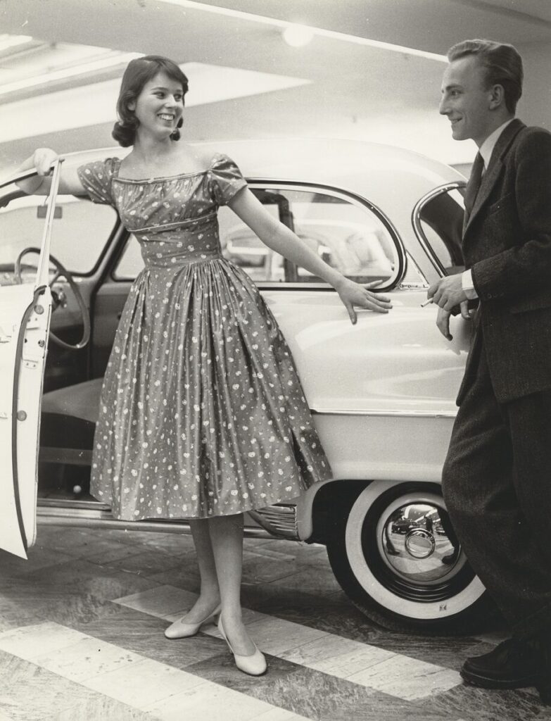 Leende ung kvinna i blommig klänning och en man i kostym med cigarett i handen. Ståendes utanför en bil.