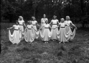 Åtta flickor med sommarklänningar som står på rad och håller ut kjolen sina klänningar, i mitten står "fröken" i förd klänning och halsband.