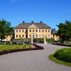 Gul herrgårdsbyggnad med grönskande park framför och grusad gångväg.