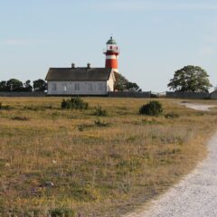 Öppet landskap med grusväg i förgrunden. I bakgrunden ett vitt hus och ett rödmålat fyrtorn.