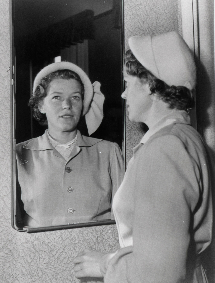 Kvinna med hatt och dräkt står och speglar överkroppen i en spegelram.
