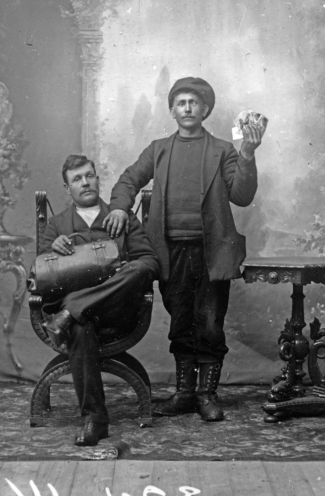 Ett något slitet gammalt foto där en man sitter med en väska i famnen och en man står bredvid med en kortlek i handen.