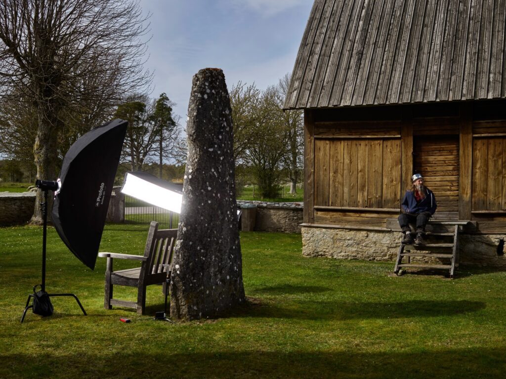 En man med skägg och mössa sitter vid en gammal fornminnesbyggnad. Bredvid står en bildsten som är upplyst med en kamera och ett fotoparaply.