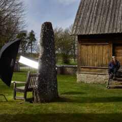 En man med skägg och mössa sitter vid en gammal fornminnesbyggnad. Bredvid står en bildsten som är upplyst med en kamera och ett fotoparaply.