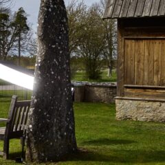 En man med skägg och mössa sitter vid en gammal fornminnesbyggnad. Bredvid står en bildsten som är upplyst med en kamera och ett fotoparaply.