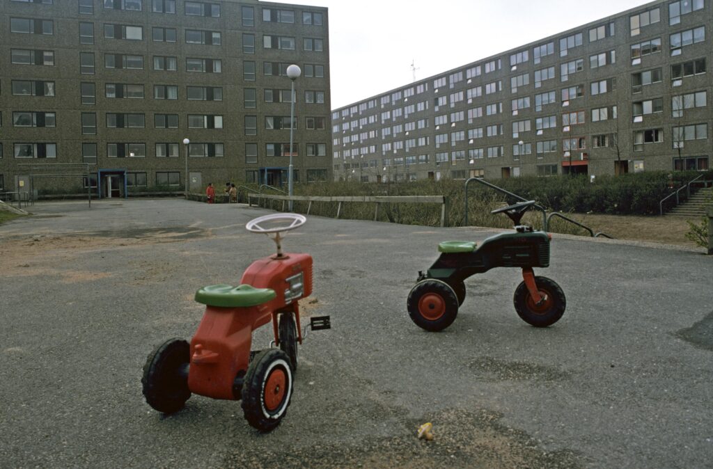 Två trehjulingar står ensamma på en asfalterad plan framför några gråa hyreshus i betong.