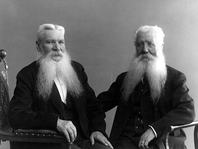 Porträtt av två vithåriga sittandes äldre män. Klädda i mörka kostymer och med likadan tvådelade skägg.