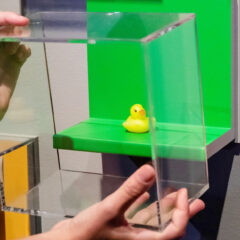 Vi ser händerna på en person som tar av skyddsglaset på en museimonter. I montern finns en gul badanka.