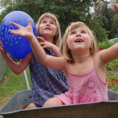 Två glada flickor sitter i en skottkärra med EHD-ballonger som lyfter mot skyn.