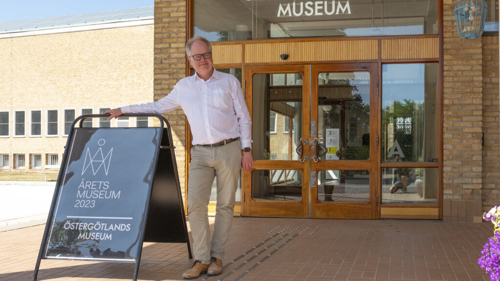 En man står utanför entrén till en museibyggnad, han lutar sig mot en skylt med texten "Årets museum 2023 Östergötlands museum".