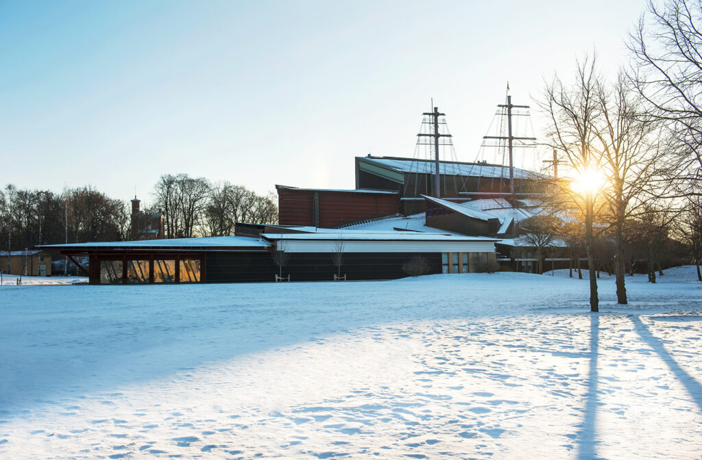 I förgrunden snötäcke och i bakgrunden museibyggnade i tre våningar med höga båtmaster i solnedgång.