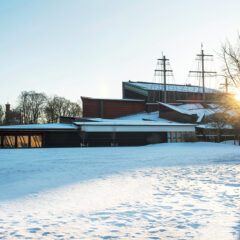 I förgrunden snötäcke och i bakgrunden museibyggnade i tre våningar med höga båtmaster i solnedgång.