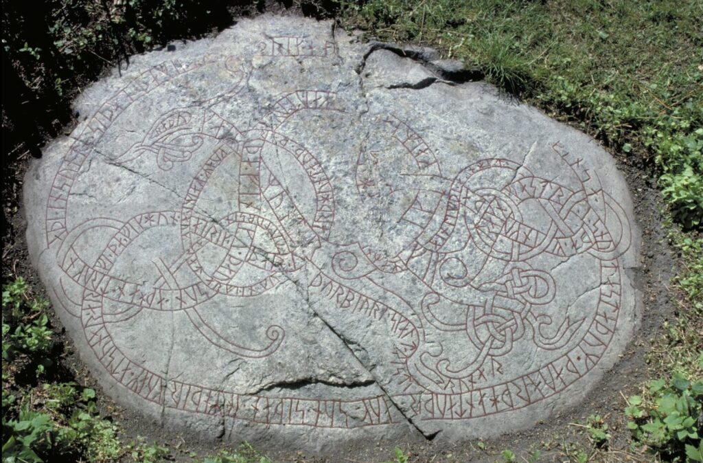 LIggande sten framgrävd i gräset. Rikt dekorerad med slungrande mönster med röda ifyllda runor.