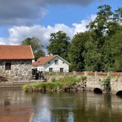 En stenbro med fyra valv ligger över en sjö, och vid änden ett gammalt hus i sten med rött tegeltak. I bakgrunden en dramatisk himmel.