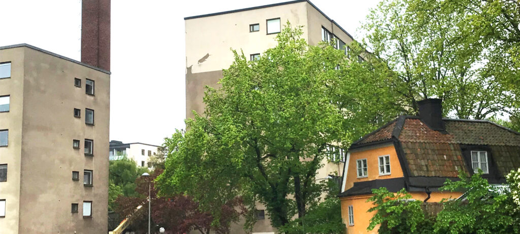Ett bostadsområde med två fyrkantiga betonghus och i förgrunden ett gult hus med brutet tegeltak omgivet av grönskande träd.
