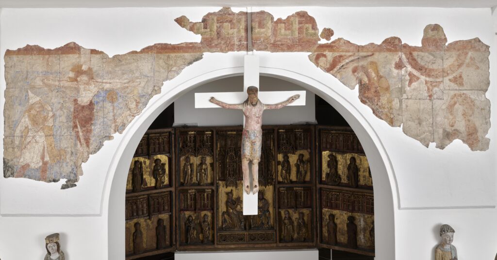 Fragment av en romansk muralmålning uppsatt på en vägg med ett valv. I mitten på bilden hänger en skulptur av Jesus på korset ner från valvet.
