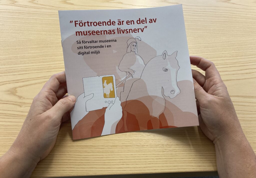 Framsidan med en tecknad bild på en historisk man sittande på en häst. Ett par händer syns hållande en mobiltelefon.