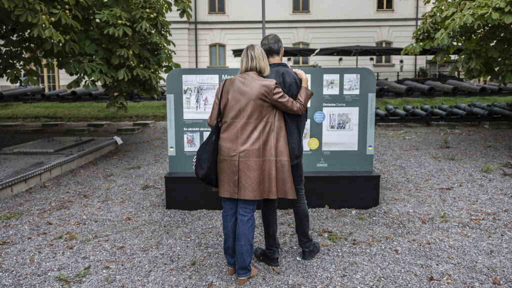 Två personer står med ryggarna mot kameran framför en utställning utanför Armémuseum.