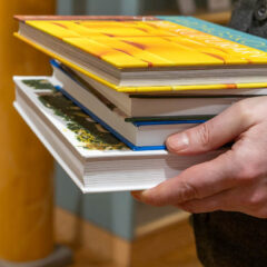 Böcker på hög som bärs av en person. Endast ena handen syns och bokhögen.