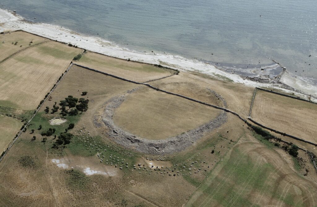 Flygbild över vy där en ringformad upphöjning av marken ligger när strand. Runtom ligger åkrar.