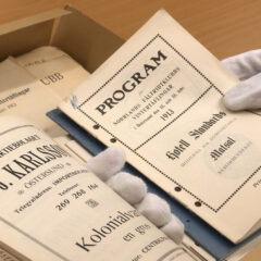 Händer med vita bomullshandskar håller i några svartvita tryckta programbroschyrer från början av 1900-talet.