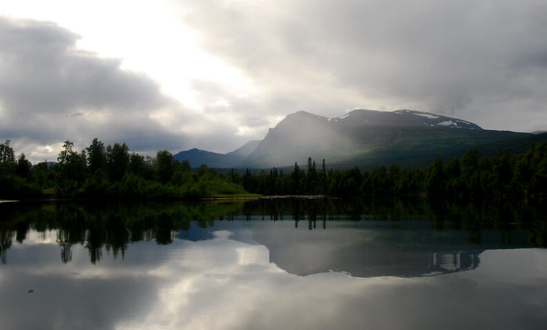 Sjö med stort bergmassiv i bakgrunden som speglas i sjön, därovan en dramatisk vacker himmel med mörka moln.