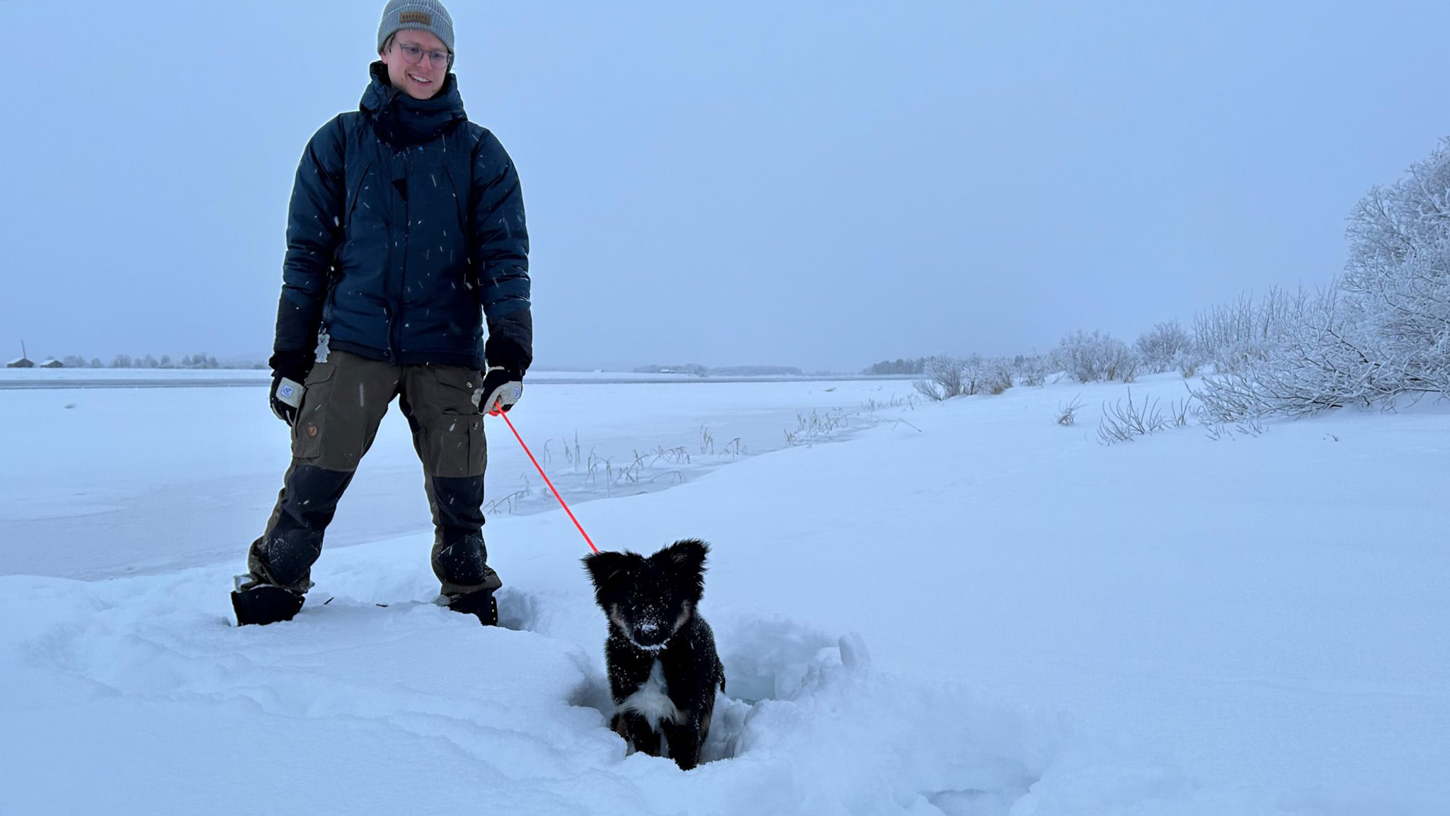 En man i vinterjacka och mössa står med en hund i koppel vid en frusen älv.