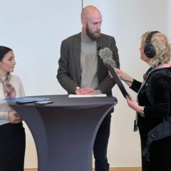 En lång man och en kort kvinna intervjuas av en reporter inomhus.