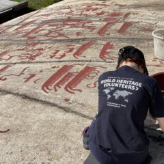 En person sitter hukad över en häll med rödmålade ristningar. Personen har en t-shirt med testen "World heritage volunteers", som talar om att det är en frivilligarbetare. Ser ut att peta bort smuts ur en skåra.