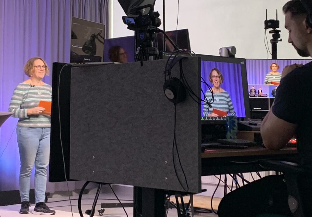 En leende kvinna står i en studio framför kameror och flera skärmar. Bakom skärmarna sitter en man och överser sändningen.