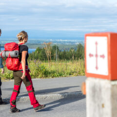Två personer ses bakifrån gåendes på en väg. De bär friluftskläder och stora ryggsäckar på ryggen. i bakgrunden skymtar man havet.