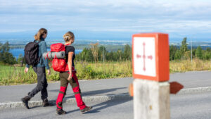 Två personer ses bakifrån gåendes på en väg. De bär friluftskläder och stora ryggsäckar på ryggen. i bakgrunden skymtar man havet.