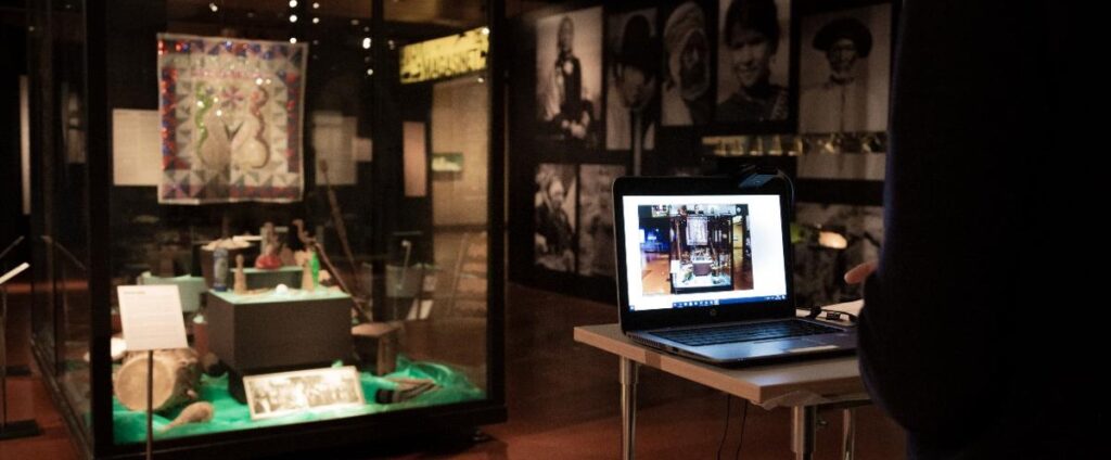 En svartklädd man står framför en museimonter med föremål. Framför sig har han en dator och på skärmen visas samma monter.