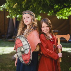 Två barn med långt hår och iklädda vikingakläder med sköld och yxa, de står rygg mot rygg,