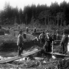 Ett röjt skogslandskap där kvinnor, män och barn står på utlagda plankor över marken som är lerig och blöt.